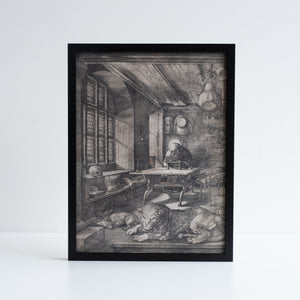 Durer etching of Saint Jerome in his Study. An etching of saint sat on a desk in a study, with dogs sleeping in front. Photographed in a black frame against a white background.