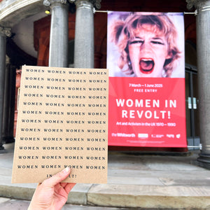 A person holding a Women in Revolt exhibition catalogue in front of the banner outside the Whitworth