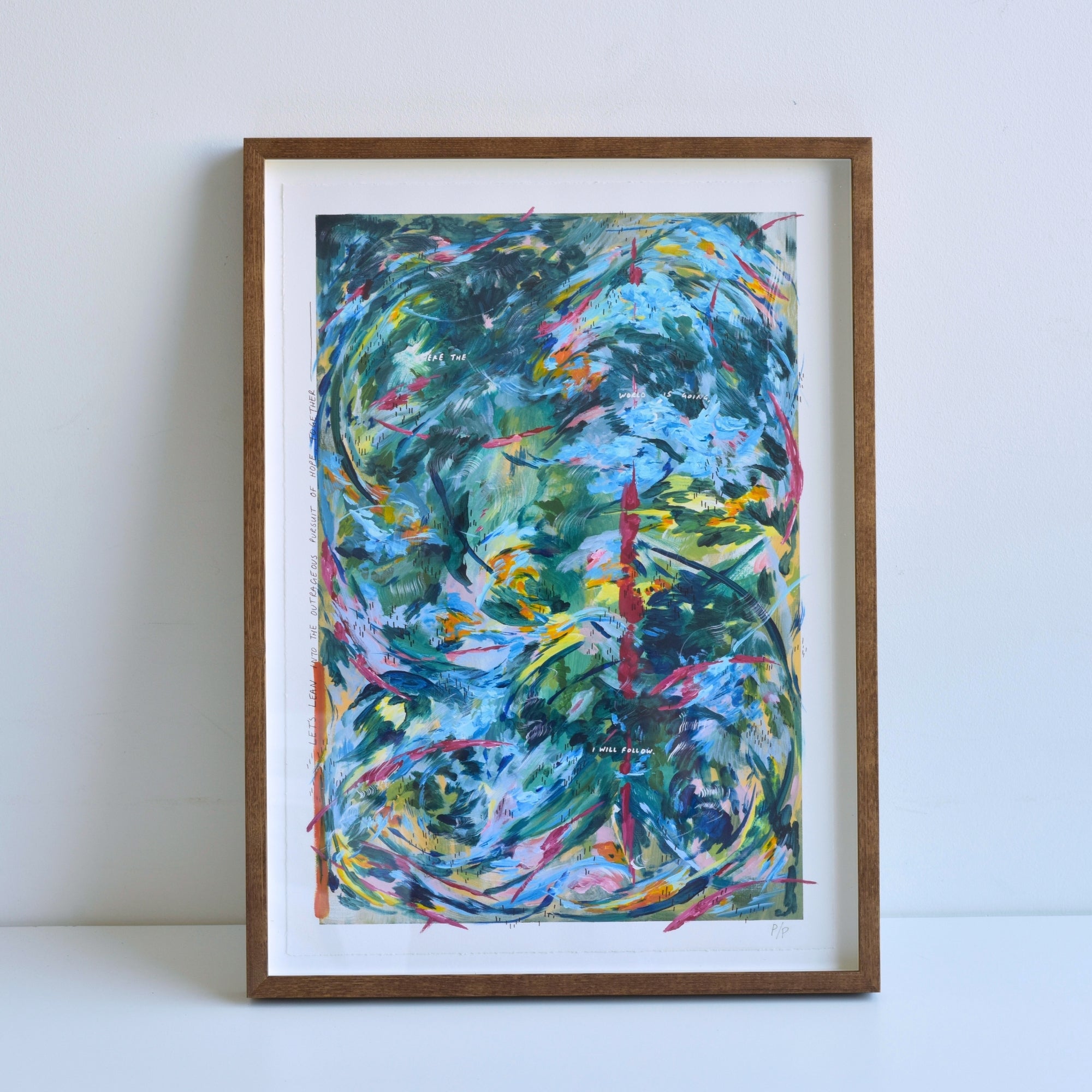 A framed abstract art print featuring swirling, expressive brushstrokes in shades of blue, green, yellow, pink, and orange, seen in a light brown wooden frame leaning against a plain white wall.