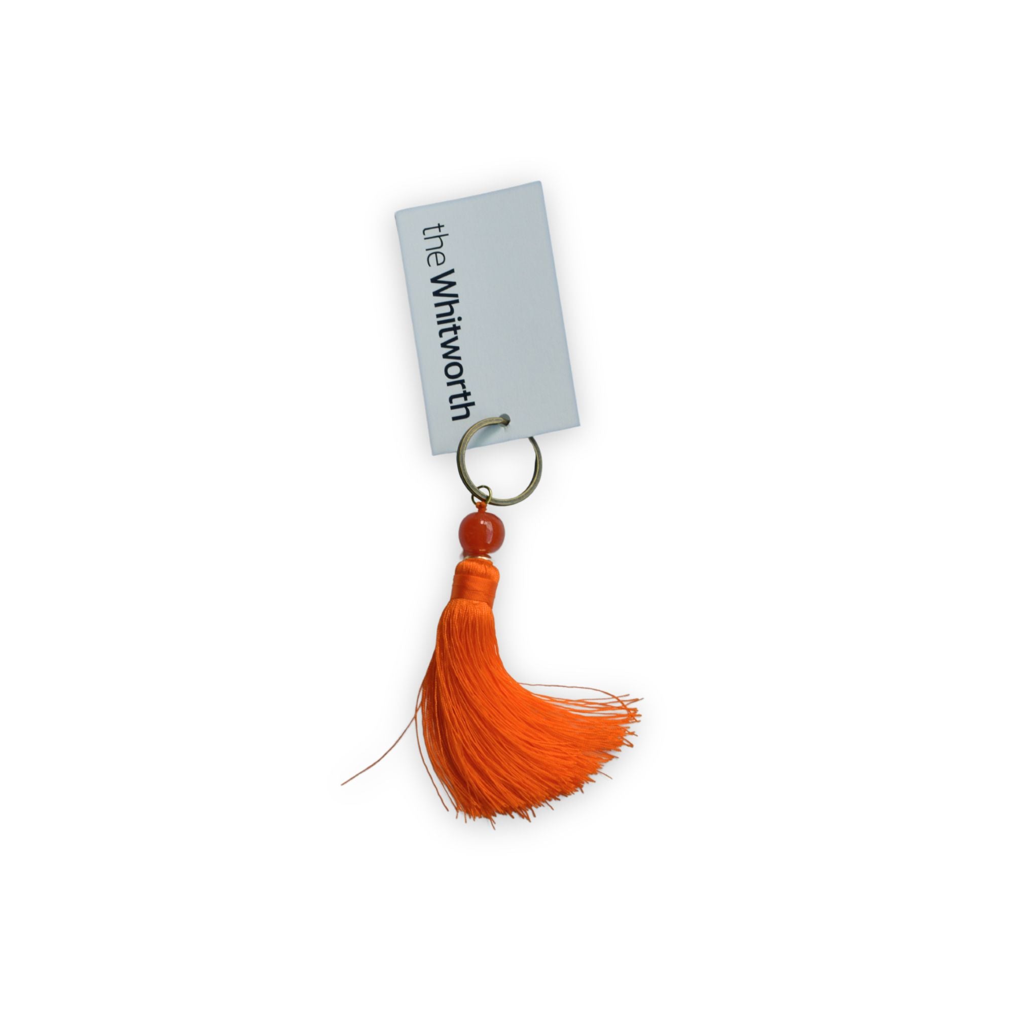 An orange tassel keychain with an orange bead and a brass key ring, attached to a swing tag printed with 'the Whitworth'.