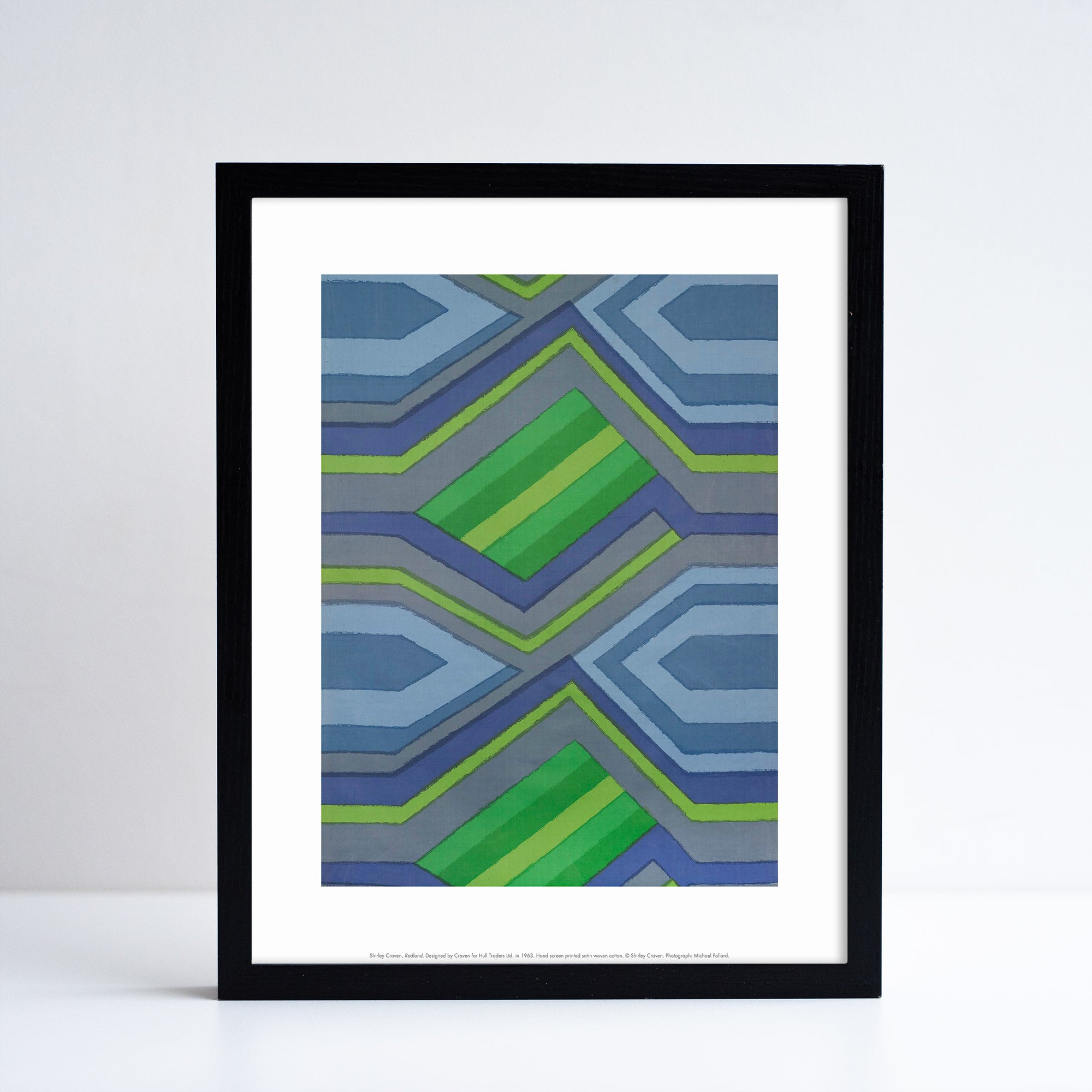 Reproduction of a green and blue geometric textiles piece by Shirley Craven, in print format with a white border. Placed in a black frame in front of a grey background.
