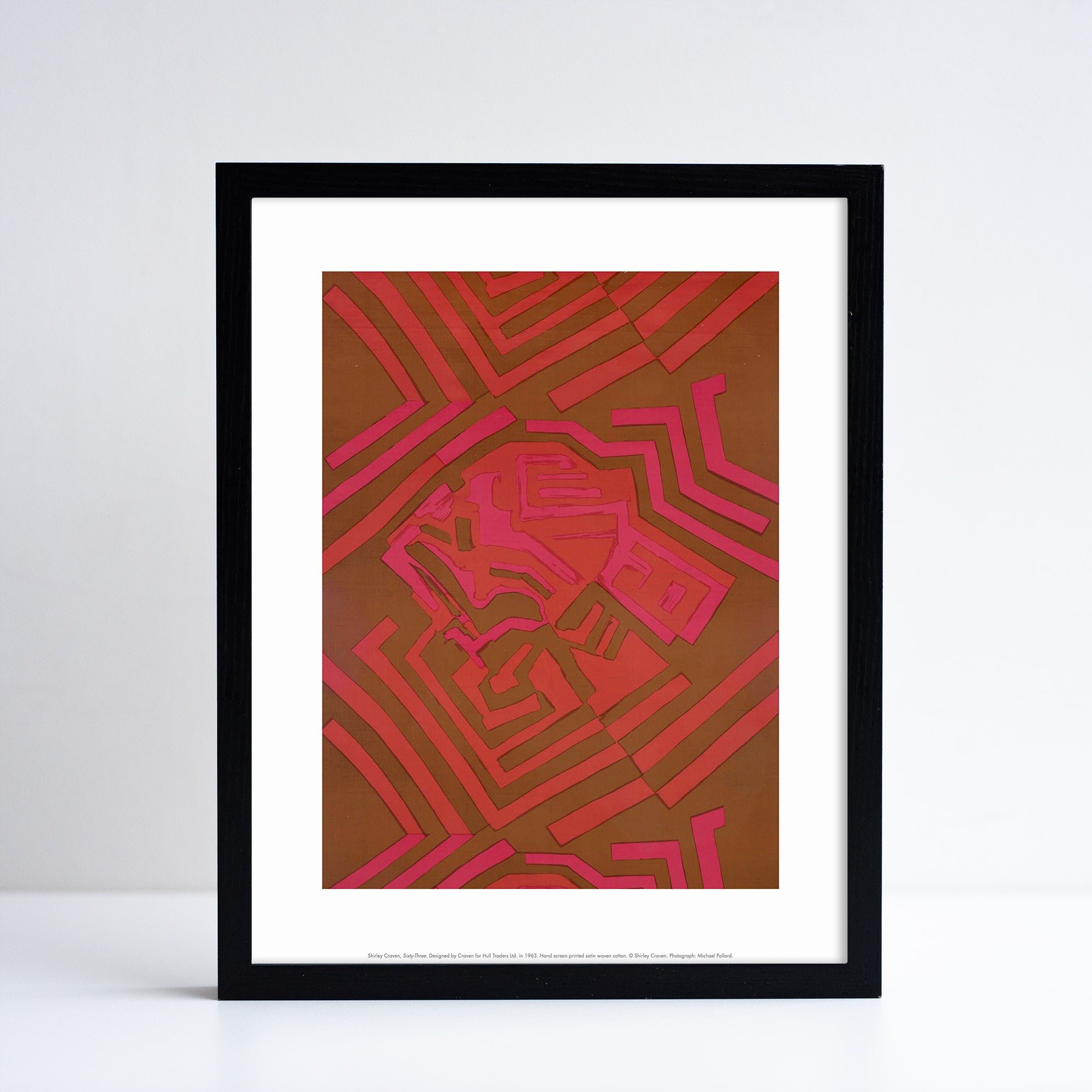 Reproduction of a red and pink geometric textiles piece by Shirley Craven, in print format with a white border. Placed in a black frame.