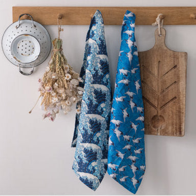 Wooden rack with kitchen items including a colander, wooden cutting board, and patterned tea towels on a white wall.