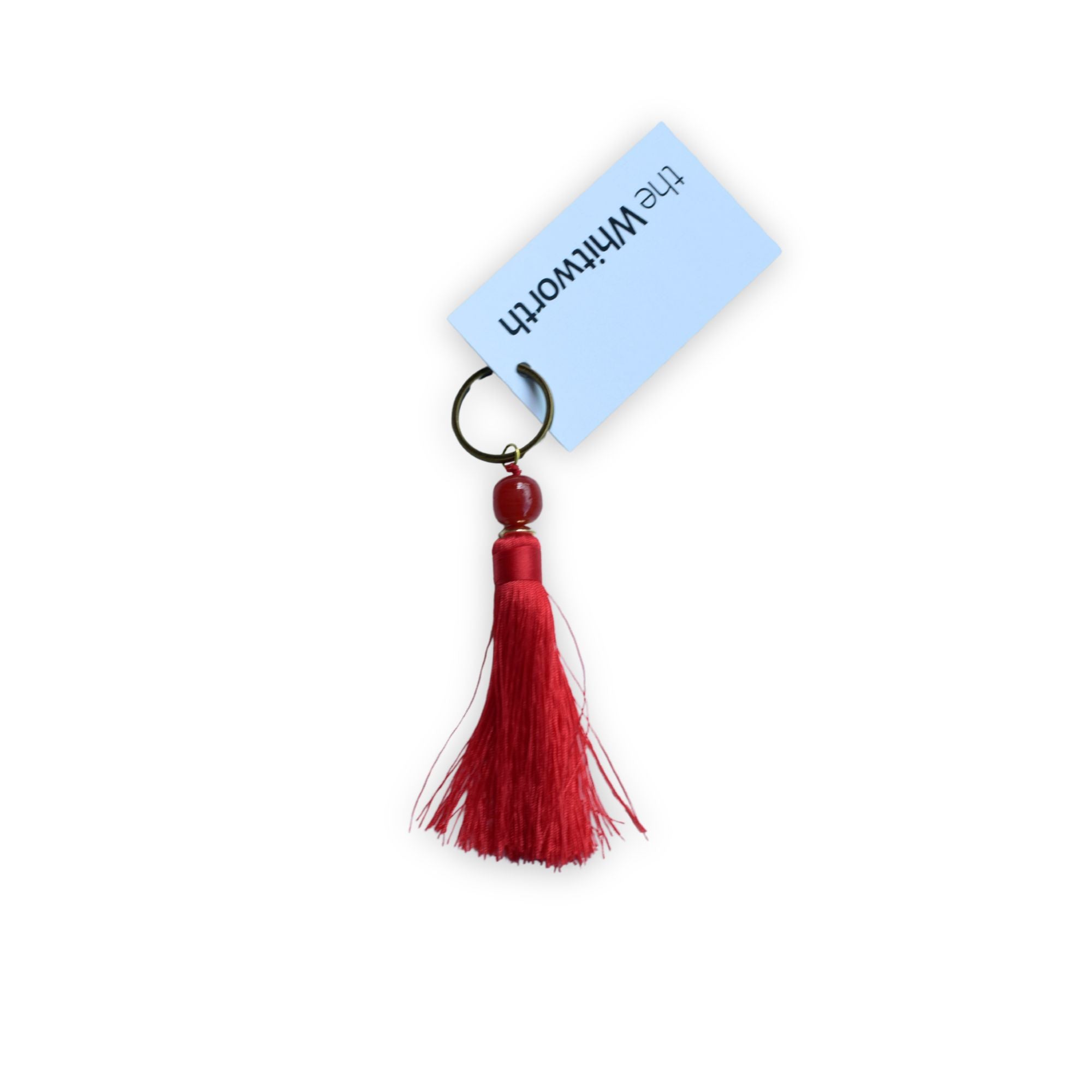 A red tassel keychain with a dark red bead and a black split ring, attached to swing tag with 'the Whitworth'.