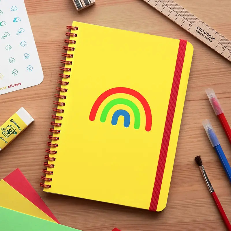 Yellow notebook with a rainbow illustration, presented against a wooden surface with stationery surrounding it.