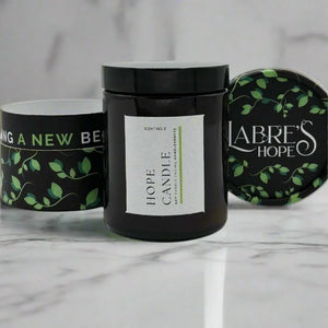 Three candles with black glass jars and white labels with green vines and the words "Hope Candle" and "Labre's Hope" sit on a white marble surface.