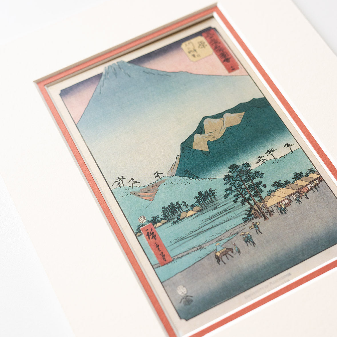 Detail a mounted artwork of a mountain landscape with a red mount on a light grey background