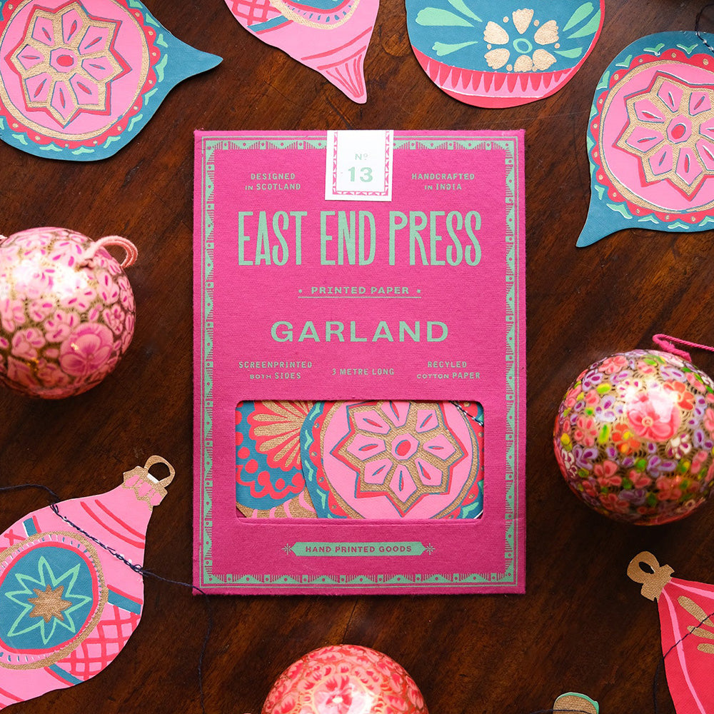 East End Press Bauble Garland in its pink paper packaging with the garland surrounding it. Photographed on a table from above.