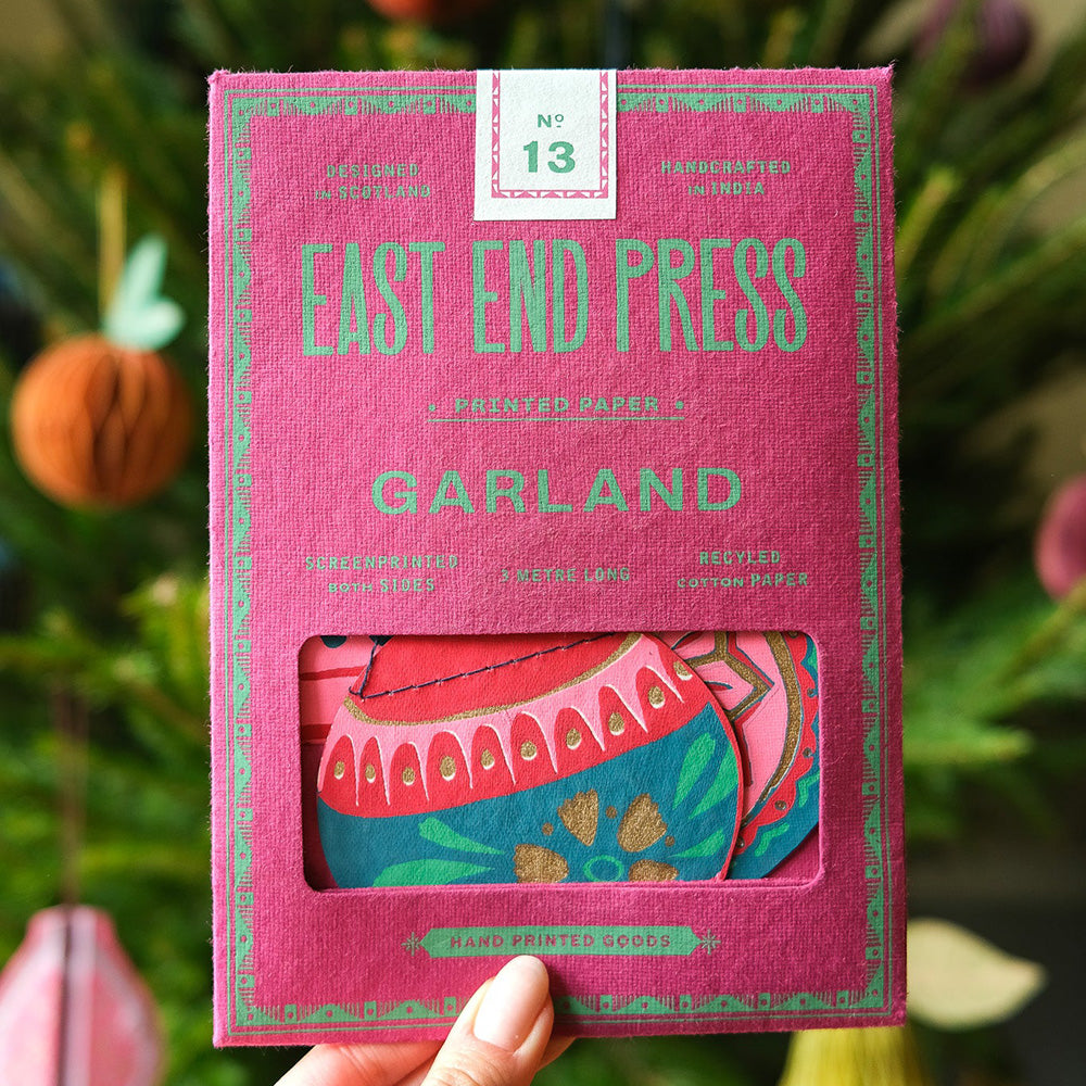 Pink packaging of East End Press Garland with floral design, held against a blurred natural background.
