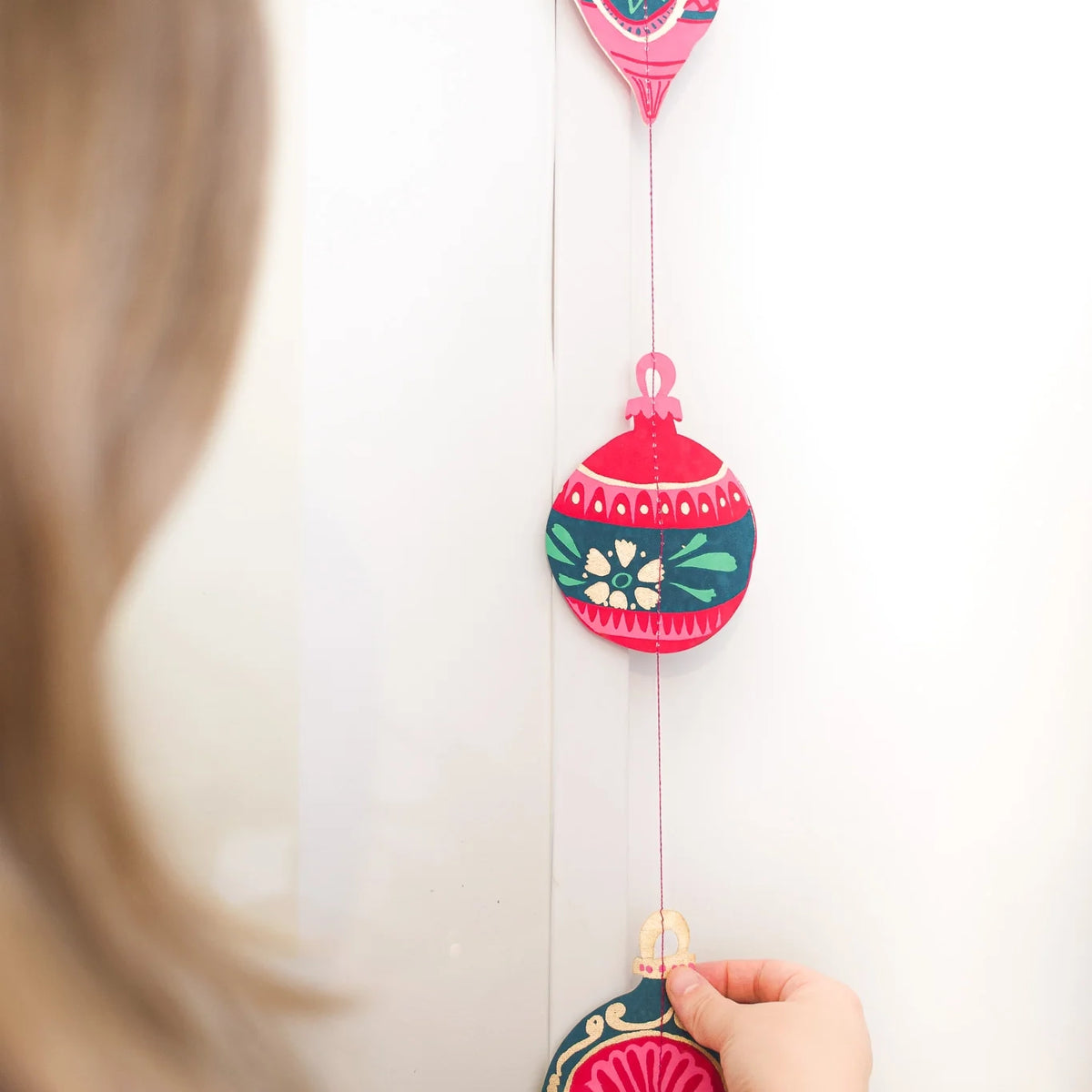 A person hanging a paper bauble decoration on a wall. The person is facing away from the camera, and their hand is visible as they attach the decoration to the string. The decoration consists of several colourful baubles with intricate patterns, connected by a thin string.