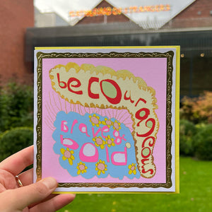 A person holding a greetings card, with yellow envelope in front of the Art Garden at The Whitworth. The card is pale pink and gold front with a blue, gold lined cloud shape at the bottom. Text is 'be courageous. brave and bold'.