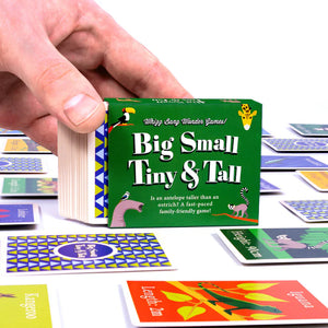 A hand holding a green card game box. The game box is surrounded by scattered cards from the game, which feature illustrations of different animals and their measurements.