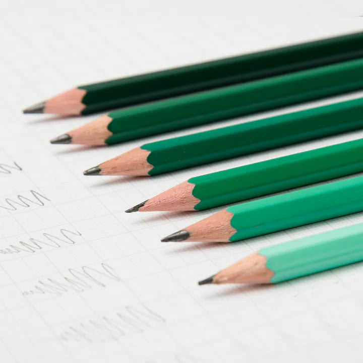 A close-up of several coloured pencils lined up on a sheet of graph paper. The pencils are in shades of green and blue.