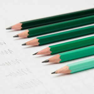 A close-up of several coloured pencils lined up on a sheet of graph paper. The pencils are in shades of green and blue.