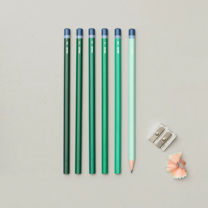 Five coloured pencils lined up in a row on a grey background. The pencils are in shades of green and blue. A pencil sharpener and pencil shavings are also on the background.