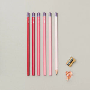 Five coloured pencils lined up in a row on a grey background. The pencils are in shades of red and pink. A pencil sharpener and pencil shavings are also on the background.