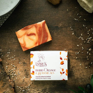 Birds-eye view of orange packaging for Labre's Hope soap, with the bar of soap placed above. Both placed on a wooden surface.