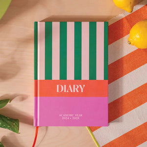 A pink and green striped diary with the text "DIARY" and "ACADEMIC YEAR 2024-2025" printed on it. The diary is lying open on a wooden surface with lemons and a plant in the background.