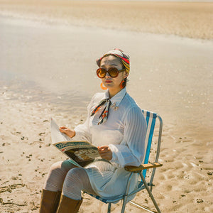 Person sitting on a beach chair reading a magazine, wearing sunglasses, the scarf and a hat.