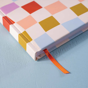 A close-up of a 2025 diary with a colourful checkered pattern cover. An orange ribbon bookmark is visible at the bottom right corner. The diary is positioned on a blue surface.