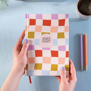 A person is holding a 2025 diary with a colourful checkered pattern cover. The diary is positioned on a blue surface with a cup of tea and pens nearby.