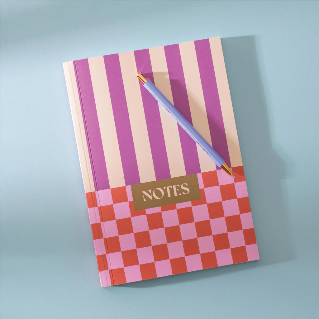 A pink and purple striped notebook with a pink and red checkered pattern on the bottom half. The word "NOTES" is written in gold lettering across the checkered section. A blue pen is resting on top of the notebook.