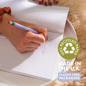 A person's hand is writing in a notebook with a purple pen. The notebook is open on a white plate, and the recycled paper symbol and labels "100% Recycled Paper," "Made in the U.K.," and "Plastic Free Packaging" are overlayed on the image.