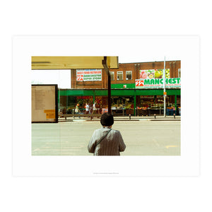 A printed reproduction of a photograph by Chris Naylor of a man looking our to a groceries shop in Manchester. 