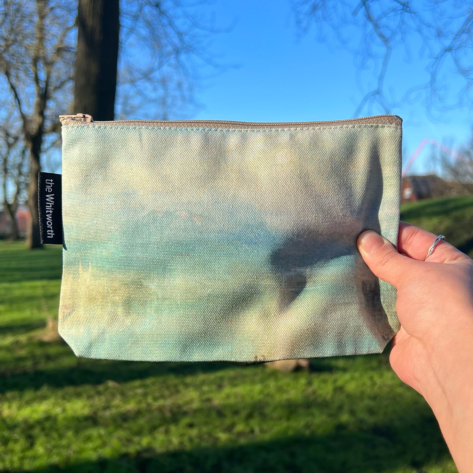 A person holding a cosmetics bag in the Whitworth Park. The bag has a reproduction of Moonlight on Lake Lucerne by William Turner.