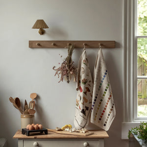 A kitchen scene with a wooden peg rail on the wall holding a dried floral bouquet, a mushroom tea towel, and a Werner's Nomenclature of Colours tea towel. Below the peg rail, a wooden countertop holds various kitchen items, including a ceramic jar of eggs, a wooden tray, and a cutting board with mushrooms and garlic.