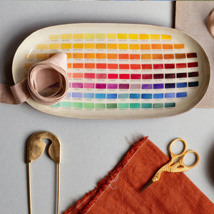 An oval-shaped enamel tray featuring a colour chart with various shades of red, orange, yellow, green, blue, and purple. The names of each colour are printed below the corresponding swatch. The tray is surrounded by other craft supplies, including a spool of ribbon, a gold safety pin, a piece of orange fabric, and a pair of gold scissors.
