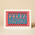 A vibrant Christmas card featuring the phrase "FELIZ NAVIDAD" in red and blue lettering, surrounded by intricate paper cut-out designs. 