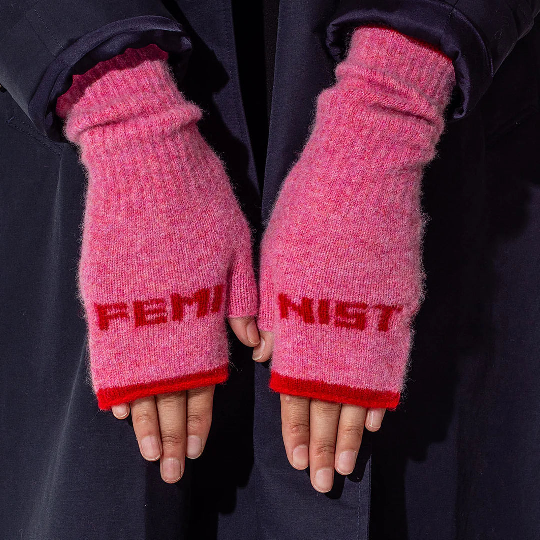 Pink fingerless gloves with 'FEMINIST' text worn by a person against a dark background