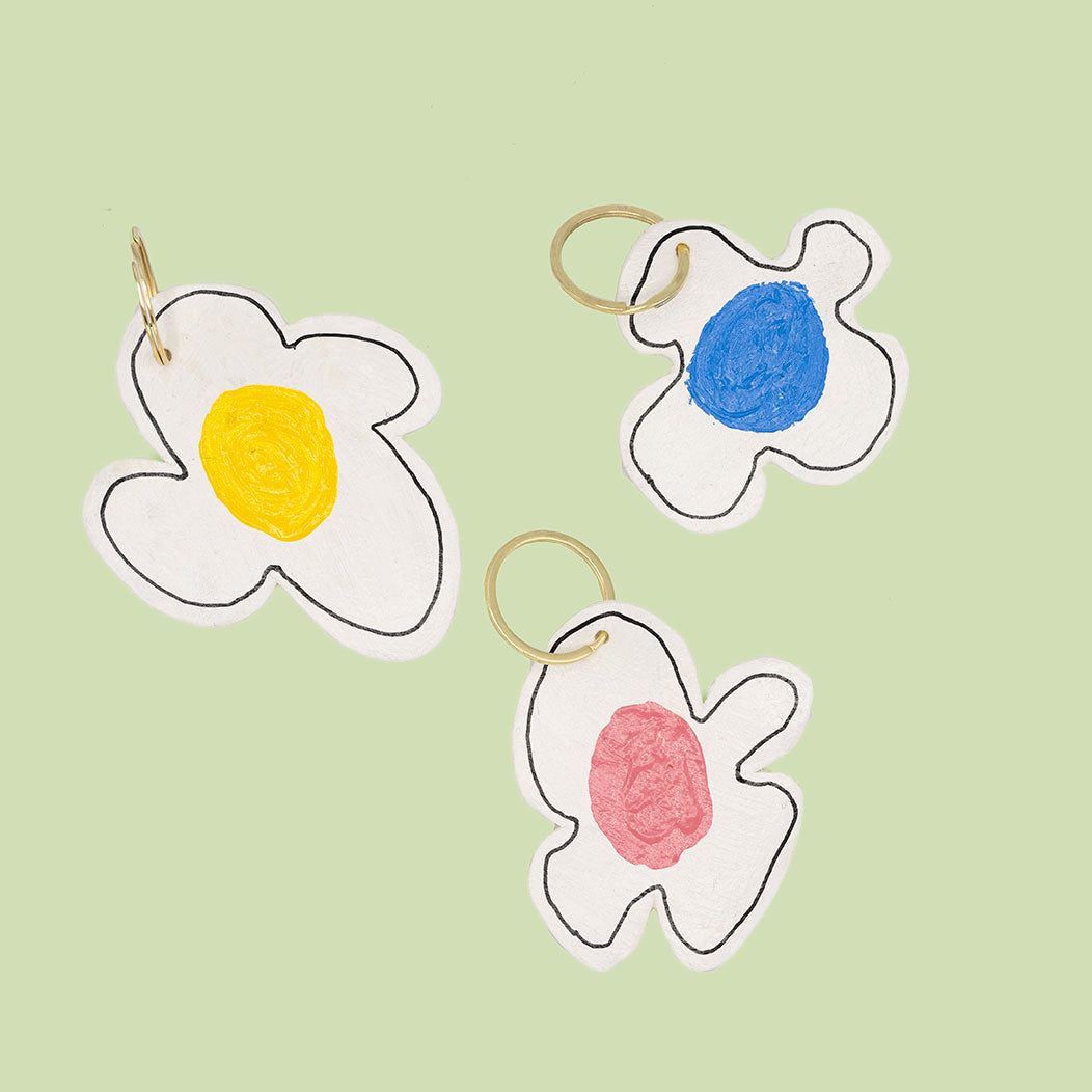 Three keyring in the shape of a flower with white petals and a varying colour centres. Photographed in front of a green background.