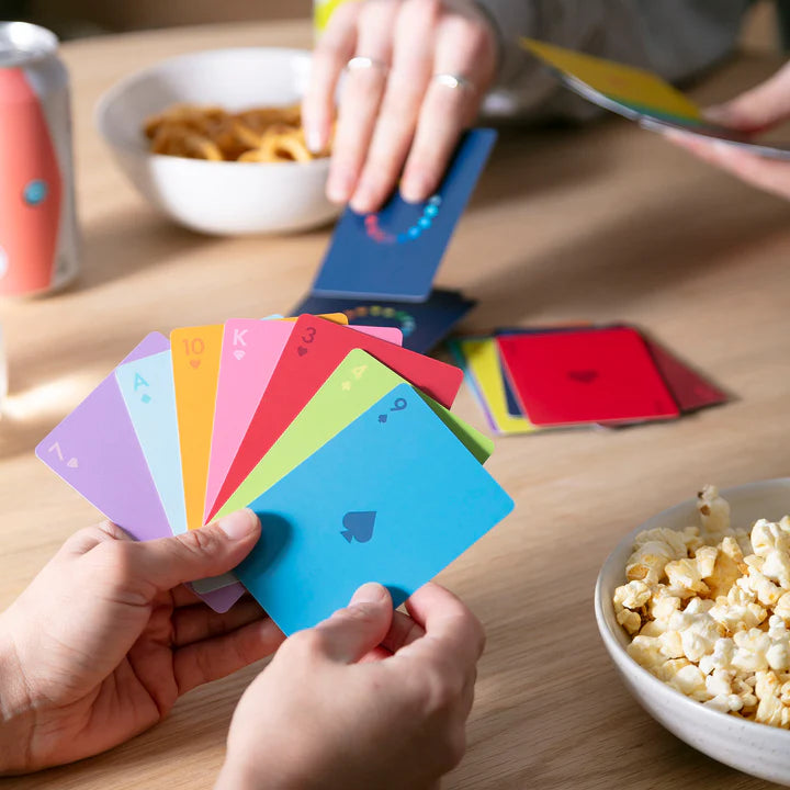 Two people playing a card game with colourful gradient playing cards. One person is holding a fan of cards, while the other is looking at their cards. There are snacks and drinks on the table.