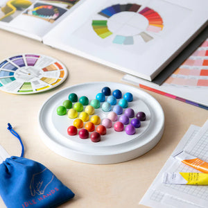 A workspace with a colour theory book, a colour wheel, paint tubes, and a circular solitaire game with colourful marbles arranged in a spiral pattern. There is also a blue pouch with the words "Idle Hands" on it.