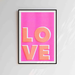 Risograph print with pink background and LOVE in bold orange letters. Placed in black frame against a grey wall.