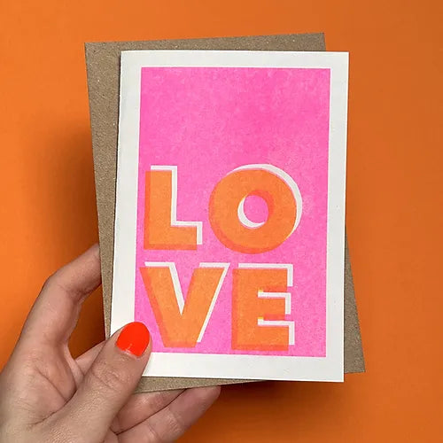 Someone holding a risograph-printed greetings card and brown envelope in front of an orange wall. The card has a pink background, white border and the word LOVE in bold orange letters at the bottom left corner of the page.