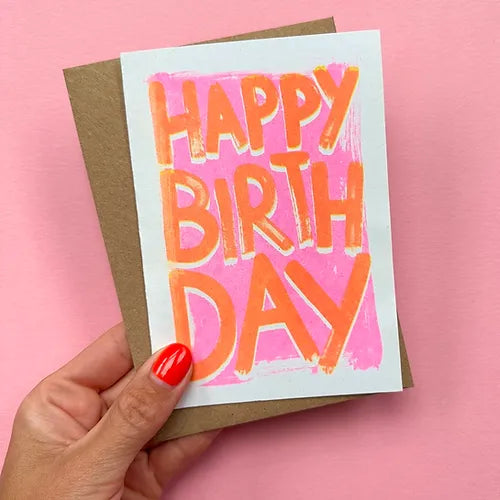 Risograph card and brown envelope held up against a pink wall. The words HAPPY BIRTHDAY are boldly laid over the card in orange lettering with a pink background.