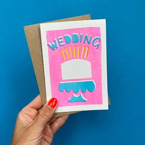 Someone holding a risograph-printed greetings and brown envelope against a blue wall. The greetings card has a pink background and stencil illustration of a cake with the word WEDDING above.