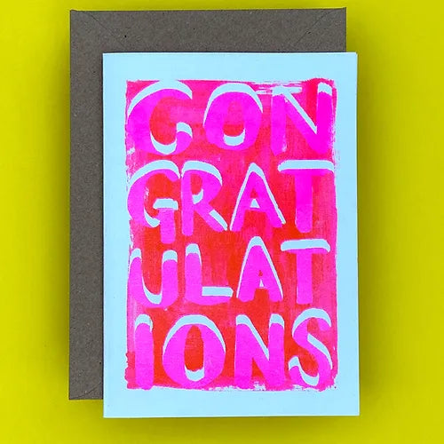 Greetings card and brown envelope on a yellow surface. The greetings card has bold pink lettering spelling 'congratulations' across the card.