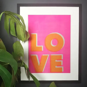 Risograph print with pink background and LOVE in bold orange letters. Placed inside a black frame with a plant next to it.