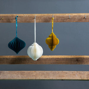 Three paper decorations in white, yellow, and teal, hanging from strings on a wooden rack against a grey wall.