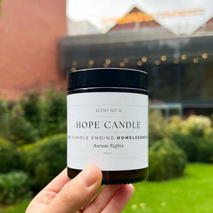 A person's hand holding a soy candle in a jar with a white label. The backdrop is the park entrance to Whitworth Art Gallery, in the art garden.