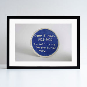 A print in a black frame. The print features a photograph of one of Horace Lindezey's ceramic 'Blue Plaques'. In this one he is describing the queen.