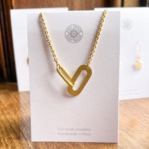 A gold necklace with an oval pendant featuring two interlocking shapes. The necklace is displayed on a card with the text.