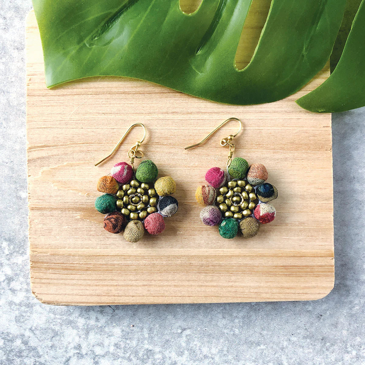 A pair of colourful beaded earrings with gold hoops, shaped like flowers. Placed ona wooden board with a plant leaf coming into the frame.