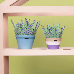 Two paper lavender plants in pots. The pots are different colours and styles, and the plants have purple flowers and green leaves. The paper plants are sitting on a shelf in front of a green wall.