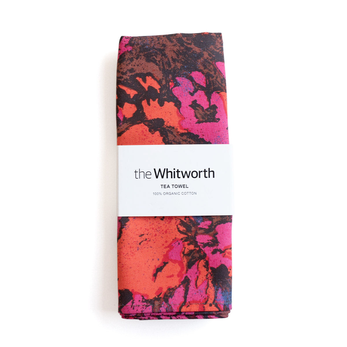 Tea towel with a vibrant pattern featuring purple and pink floral designs. This pattern is a detail of a design by Shirley Craven. Belly band around the tea towel with the Whitworth logo.