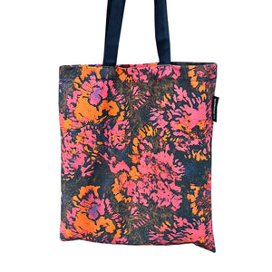 The image shows a tote bag with a vibrant floral pattern in shades of pink, orange, and black. The tote bag has black handles.